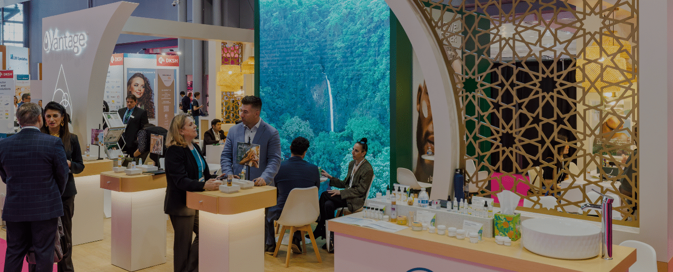 A zoomed in picture of the Vantage booth at InCosmetics the focal point being the large graphic wall portraying a forest, a range of small tables holding products where people are chatting 