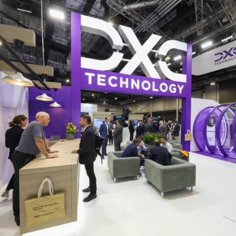 A front view of the DXC Technology stand at DSEI where the focal point is a large branded signage and a purple circular tunnel with a dart board inside where people are playing and chatting