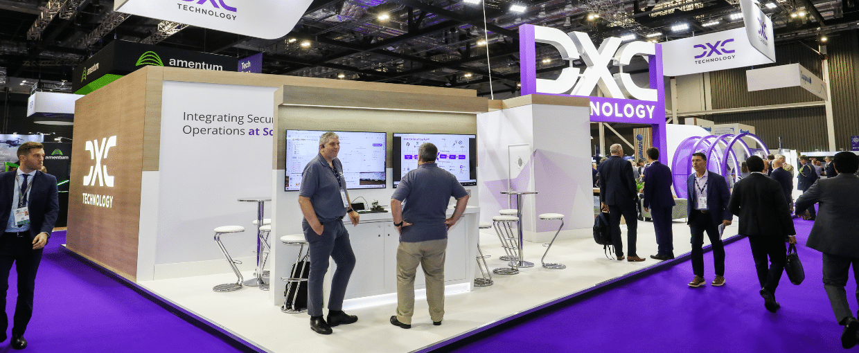 The DXC booth at DSEI with large branded signage, two tv screens where people are chatting and a range of high tables with chairs