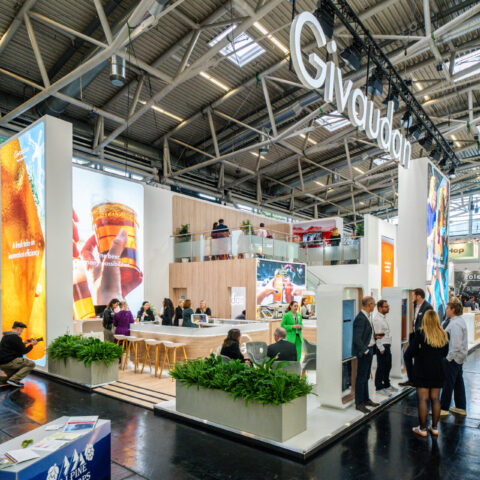 A wide view of the Givaudan booth at Drinktec where there is large branded signage above the booth and large graphics within it, a range of tables and chairs with an upstairs bar area