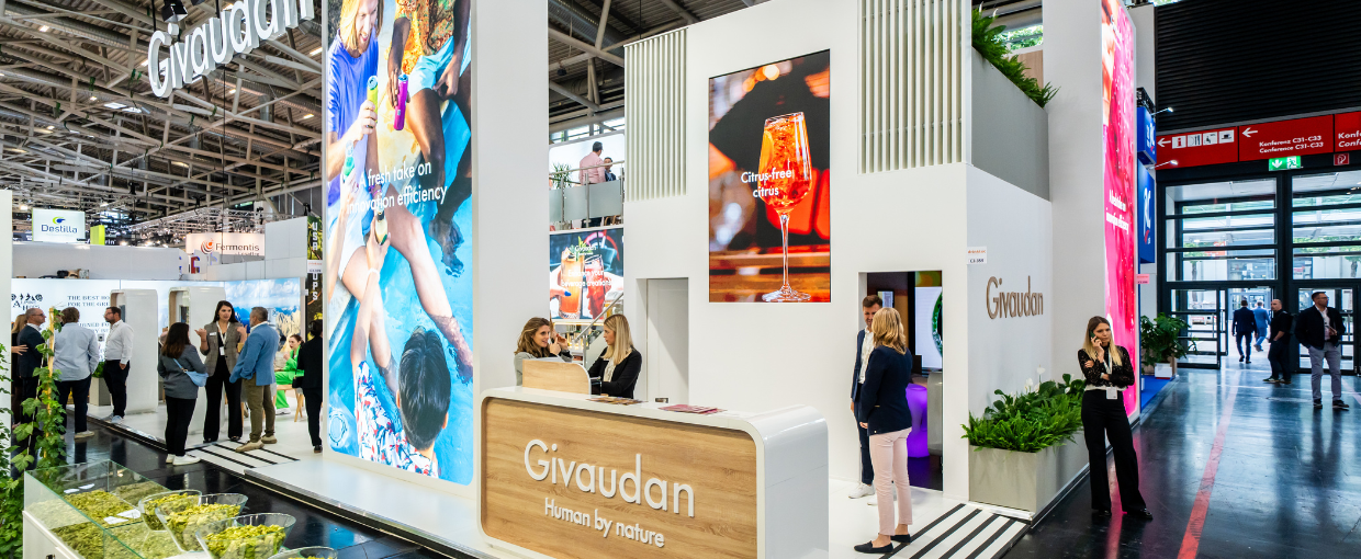A banner of the Givaudan booth at Drinktec where there is a reception counter with branded signage, large graphic rectangles and two rooms with the doors slightly open
