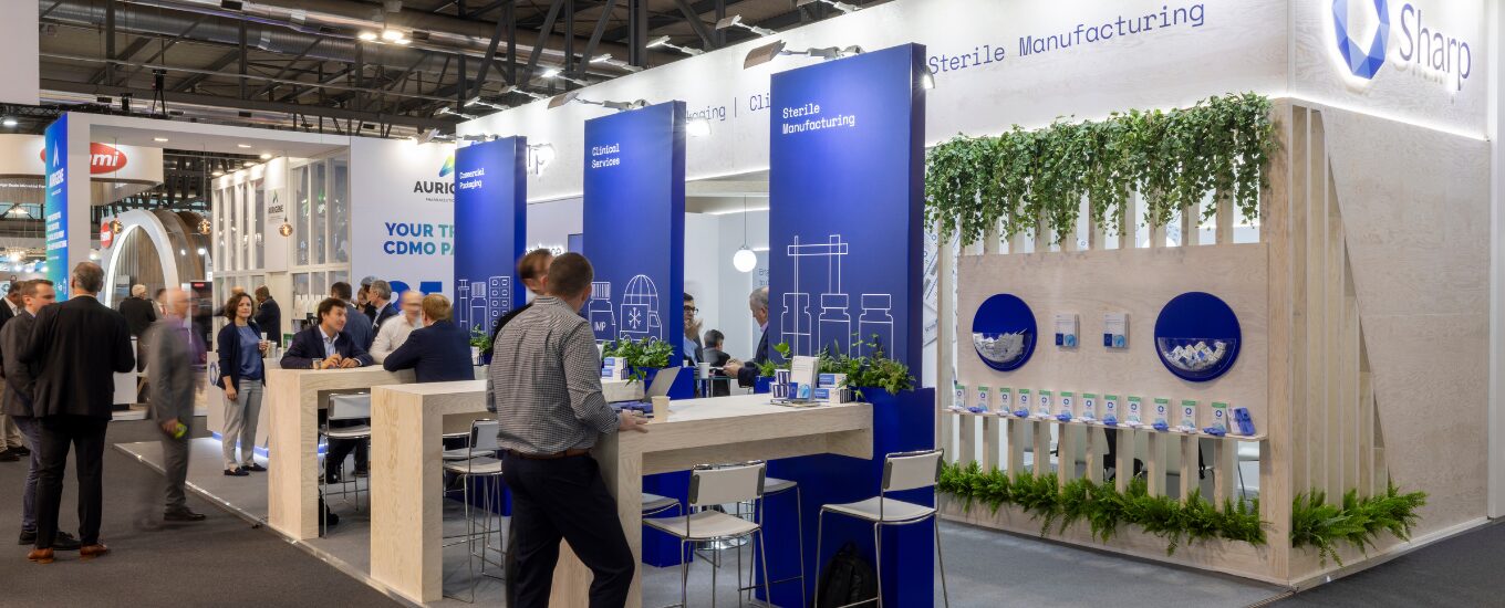 Sharp booth at CPHI 2024 angled shot of the booth with large commercial signage, meeting tables and a modern product wall and multiple visitors chatting over the meeting tables